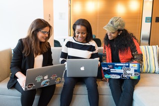 Three women huddling together on a couch with their laptops