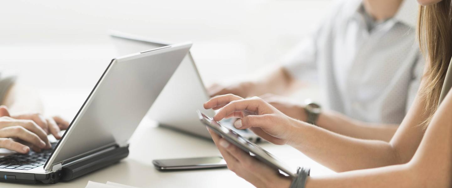 Three individuals sitting at a desk looking at their respective laptops and smartphone