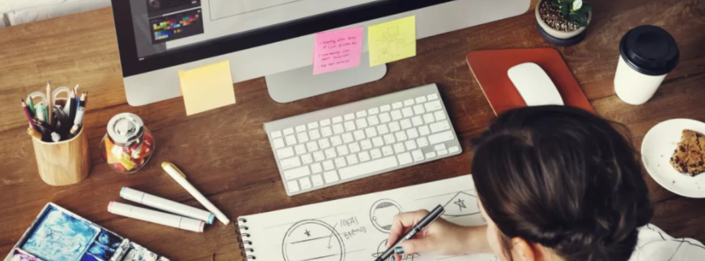 A person sketching on a pad seated in front of a computer