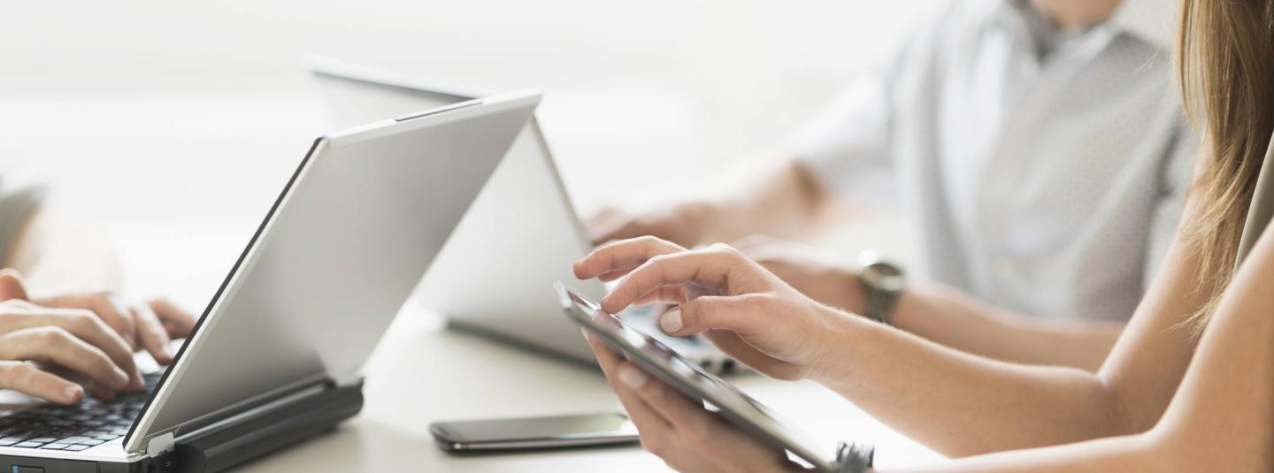 Three individuals sitting at a desk looking at their respective laptops and smartphone