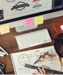 A person sketching on a pad seated in front of a computer