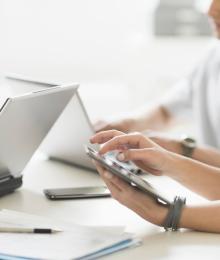 Three individuals sitting at a desk looking at their respective laptops and smartphone